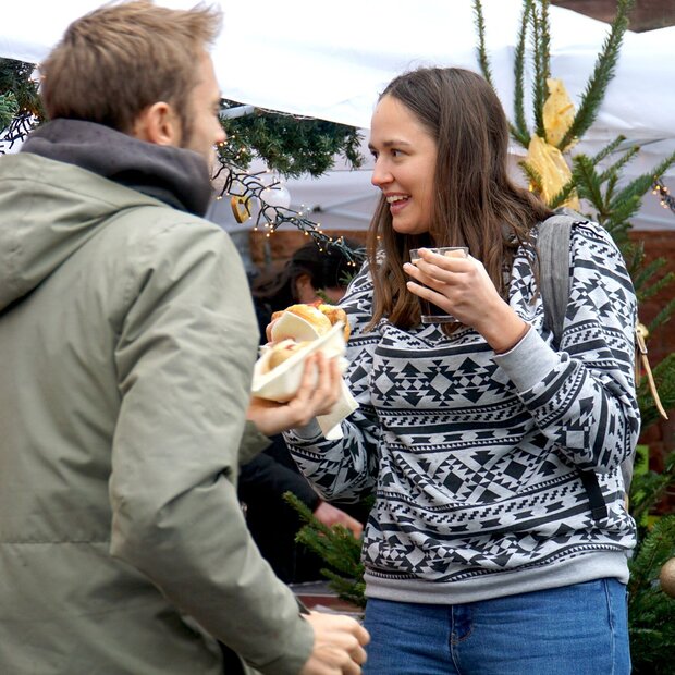 Eine junge Frau steht an einem Weihnachtsstand der Pädagogischen Hochschule Heidelberg. Sie lächelt herzlich und hält etwas zu Essen sowie Trinken in der Hand. Von schräg hinten sieht man einen jungen Mann, der etwas zu Essen in der Hand hält.