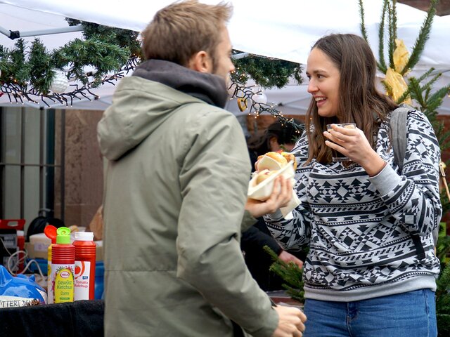 Eine junge Frau steht an einem Weihnachtsstand der Pädagogischen Hochschule Heidelberg. Sie lächelt herzlich und hält etwas zu Essen sowie Trinken in der Hand. Von schräg hinten sieht man einen jungen Mann, der etwas zu Essen in der Hand hält.