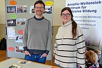 Das Bild zeigt Florian Kollmann und Anna Neff vor einem Tisch am Markt der Möglichkeiten. Hinter ihnen sind verschiedene Stell- und Pinnwände. Beide lächeln in die Kamera.