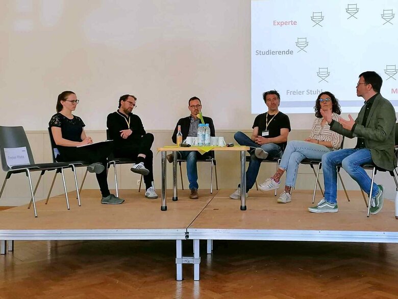 Sechs Personen sitzen auf einem Podium im Halbkreis; ein siebter Platz links ist leer. Der Mann rechts spricht deutlich mit den Händen. Die anderen Personen hören ihm zu. Das Bild wurde in der Aula der Pädagogischen Hochschule während einer Veranstaltung zu herausforderndem Verhalten aufgenommen.