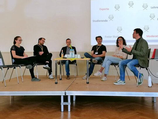 Sechs Personen sitzen auf einem Podium im Halbkreis; ein siebter Platz links ist leer. Der Mann rechts spricht deutlich mit den Händen. Die anderen Personen hören ihm zu. Das Bild wurde in der Aula der Pädagogischen Hochschule während einer Veranstaltung zu herausforderndem Verhalten aufgenommen.