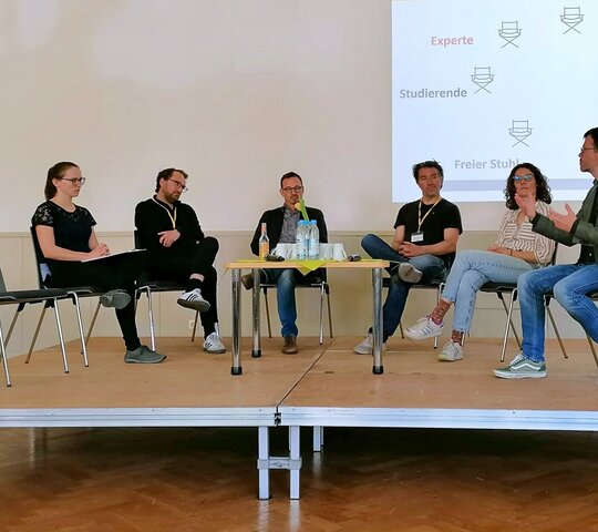 Sechs Personen sitzen auf einem Podium im Halbkreis; ein siebter Platz links ist leer. Der Mann rechts spricht deutlich mit den Händen. Die anderen Personen hören ihm zu. Das Bild wurde in der Aula der Pädagogischen Hochschule während einer Veranstaltung zu herausforderndem Verhalten aufgenommen.