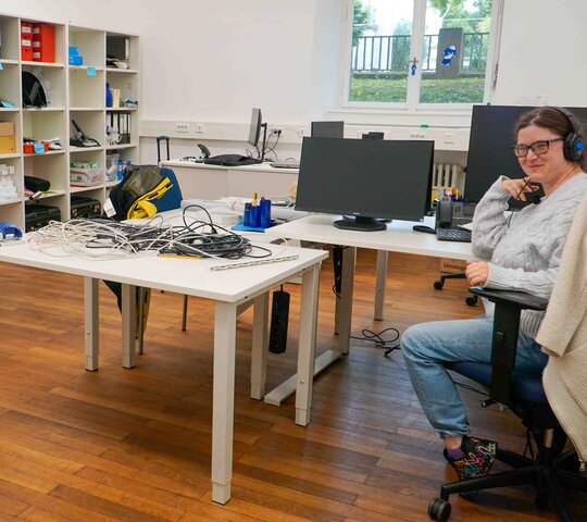 Das Bild zeigt ein Büro des AW-ZIB. Zwei Frauen sitzen am Rand an Schreibtischen, eine lächelt in die Kamera, eine schaut nach unten und arbeitet. Im Raum verteilt stehen Tische, auf denen viele Kabel liegen.
