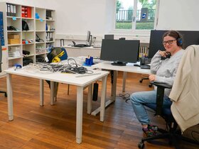 Das Bild zeigt ein Büro des AW-ZIB. Zwei Frauen sitzen am Rand an Schreibtischen, eine lächelt in die Kamera, eine schaut nach unten und arbeitet. Im Raum verteilt stehen Tische, auf denen viele Kabel liegen.