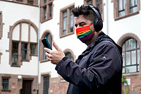 Auf dem Foto ist Klemens zu sehen. Er steht von der PH Heidelberg. Klemens trägt Kopfhörer und schaut auf sein Handy. Er trägt zudem einen Mund-Nase-Schutz in Regenbogenfarben.