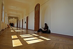 Das Bild zeigt einen Gang des Altbaus der Ph mit einer Studentin die vor einer Tür sitzt.