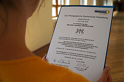 Das Bild fokussiert auf die Verleihungsurkunde, die von einer Frau gehalten wird. Das Foto wurde im Altbau der Hochschule aufgenommen. Copyright Pädagogische Hochschule Heidelberg
