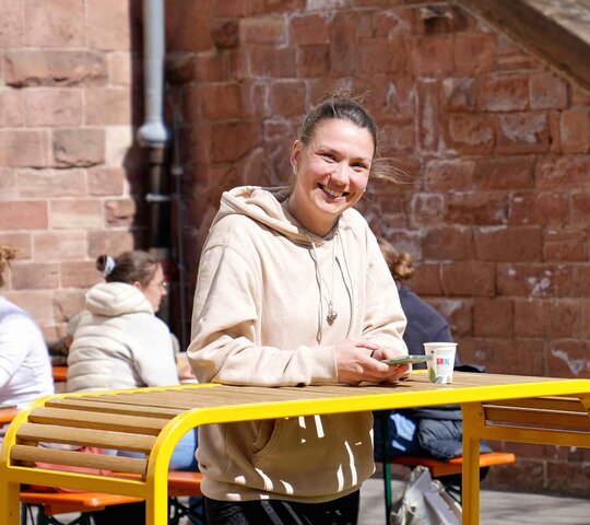Eine junge Frau steht im Innenhof der Hochschule an einem Stehtisch und strahlt direkt in die Kamera.