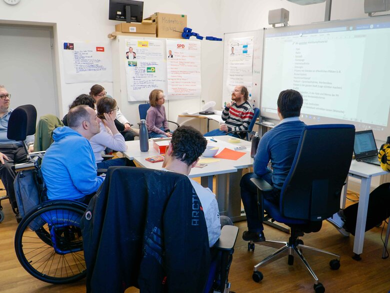 Bildungsfachkräfte und Qualifizierungsteilnehmende sitzen im Qualifizierungsbüro im Kreis und diskutieren. Vor ihnen ist ein Whiteboard, an den Wänden hängen FlipCharts, die die Positionen einiger Parteien zusammenfassen.
