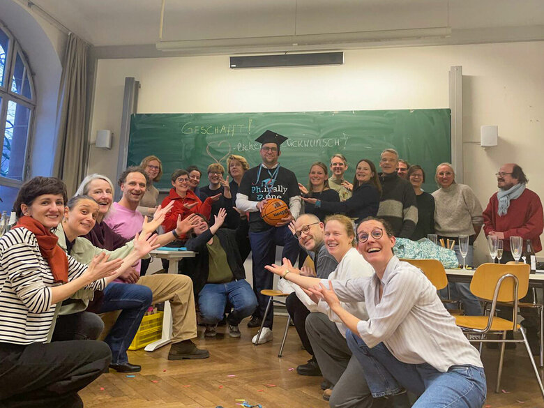 Das Bild zeigt David Dörrer, nach seiner erfolgreichen Disputation. Er steht in der Mitte eine Seminarraums, um ihn herum knien und stehen Freunde, Familienmitglieder und ein Teil des AW-ZIB-Teams. David Dörrer hält einen Basketball in der Hand und trägt auf dem Kopf einen Doktorhut.