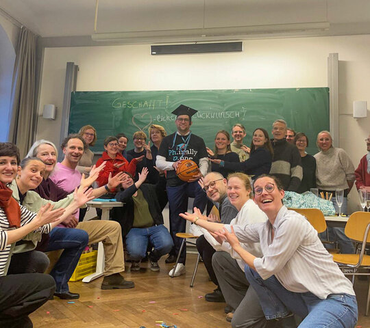 Das Bild zeigt David Dörrer, nach seiner erfolgreichen Disputation. Er steht in der Mitte eine Seminarraums, um ihn herum knien und stehen Freunde, Familienmitglieder und ein Teil des AW-ZIB-Teams. David Dörrer hält einen Basketball in der Hand und trägt auf dem Kopf einen Doktorhut.