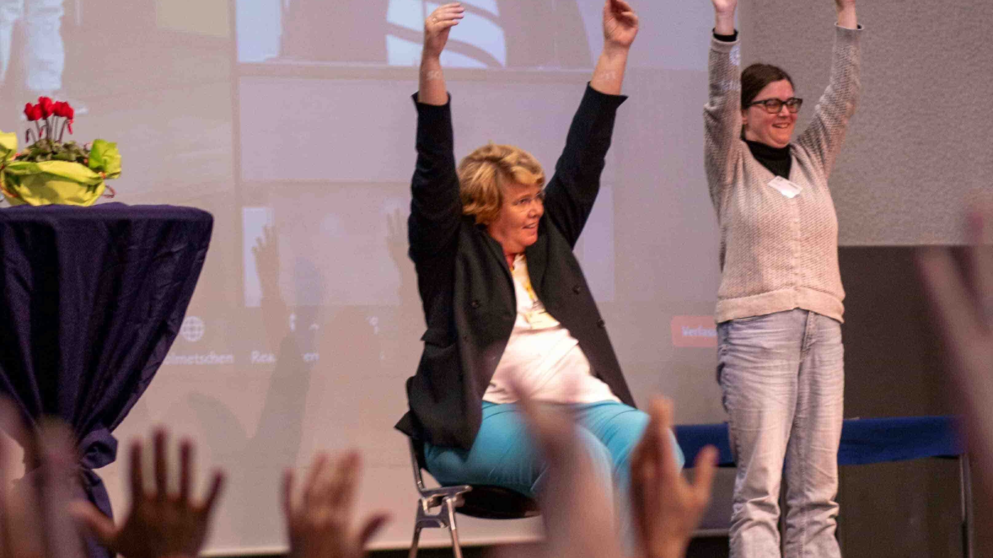Prof. Dr. Karin Terfloth und Bildungsfachkraft Anna Neff sind auf der Bühne. Sie machen Übungen der bewegten Pause vor und strecken die Arme in die Luft.