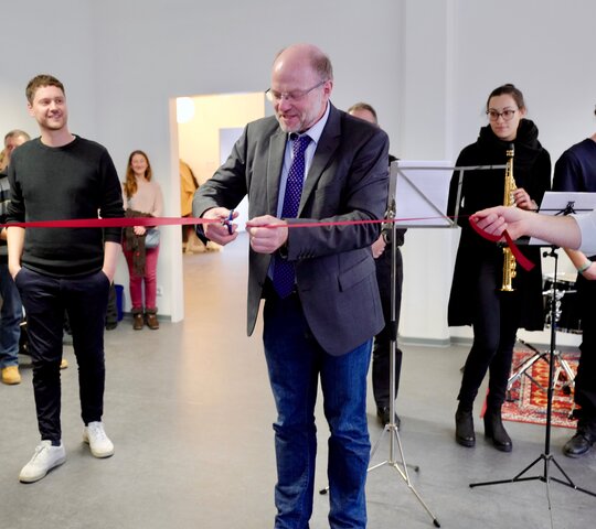 Zu sehen sind mehrere Personen bei einer Feierlichkeit. Eine Person durchschneidet ein rotes Band.