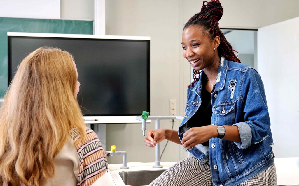 Eine junge Frau sitzt auf einem Tisch. Sie spricht deutlich mit den Händen zu einer anderen Frau, die man im linken Bildausschnitt von hinten sieht. Das Bild wurde im Biologielabor der Pädagogischen Hochschule Heidelberg aufgenommen.