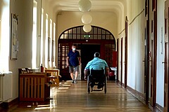 Das Bild zeigt einen Mann im Rollstuhl von hinten, welcher einen Flur der Pädagogischen Hochschule Heidelberg hinaufrollt. Diesem kommt dabei ein gehender Mann mit Mundschutz entgegen.