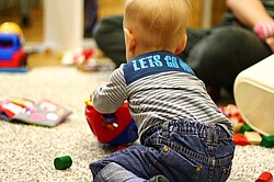 Kleinkind von hinten, welches mit Spielzeug auf einem Teppich spielt. 