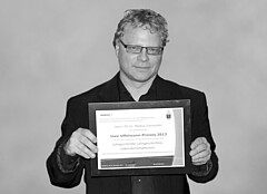 Die Poträutaufnahme zeigt Markus Daumüller. Auf dem Schwarz-Weiß Bild hält er die Urkunde des Uwe-Uffelmann-Preises in die Kamera.