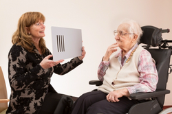 Auf dem Bild ist links eine Frau zu sehen, die eine weiße Tafel mit vier schwarzen Strichen hoch hält. Rechts sieht man eine alte Dame, die im Rollstuhl sitzt. Sie schaut nachdenklich auf die Tafel. Copyright: Blindeninstitut/Modellprojekt „Sehen im Alter“