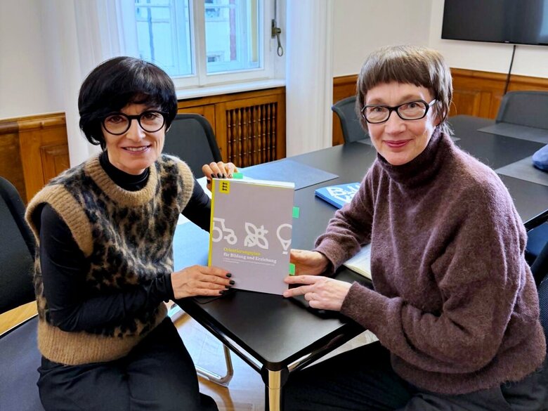 Bürgermeistern Jansen und Rektorin Vach sitzen an einem Tisch und halten den „Orientierungsplan für Bildung und Erziehung in Kindertageseinrichtungen“ zwischen sich. Beide schauen freundlich in die Kamera.