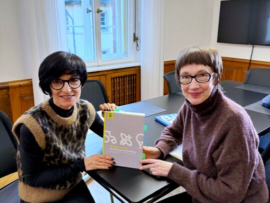 Bürgermeistern Jansen und Rektorin Vach sitzen an einem Tisch und halten den „Orientierungsplan für Bildung und Erziehung in Kindertageseinrichtungen“ zwischen sich. Beide schauen freundlich in die Kamera.