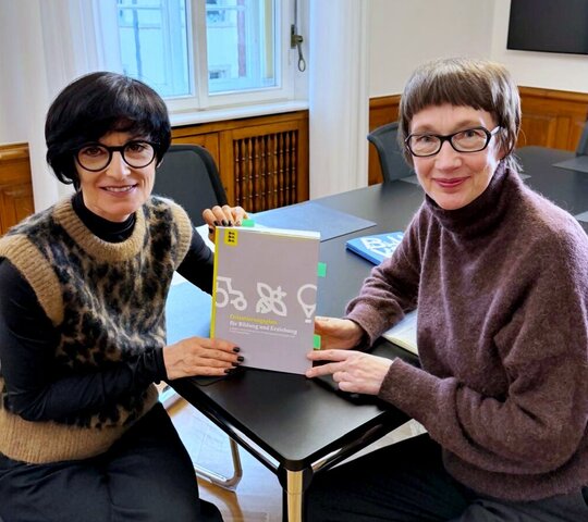 Bürgermeistern Jansen und Rektorin Vach sitzen an einem Tisch und halten den „Orientierungsplan für Bildung und Erziehung in Kindertageseinrichtungen“ zwischen sich. Beide schauen freundlich in die Kamera.