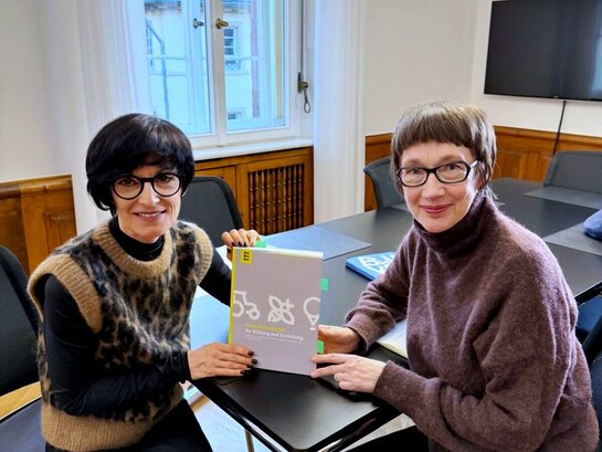 Bürgermeistern Jansen und Rektorin Vach sitzen an einem Tisch und halten den „Orientierungsplan für Bildung und Erziehung in Kindertageseinrichtungen“ zwischen sich. Beide schauen freundlich in die Kamera.