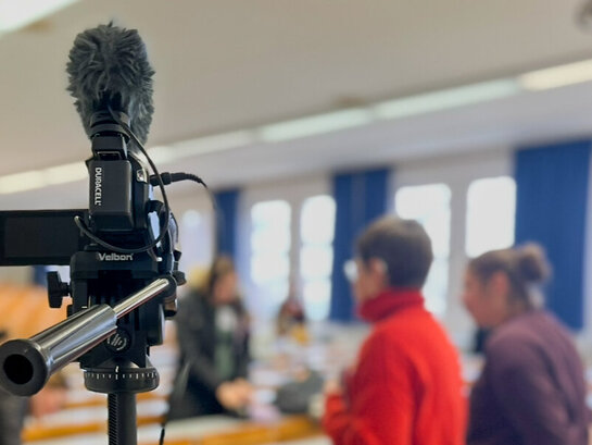 Nahaufnahme eines Stativs mit Kamera, im Hintergrund sind verschwommen Personen in einem Hörsaal