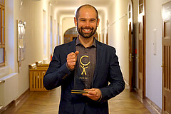Das Bild zeigt Sebastian Stumpf. Er steht im Altbauflur der Hochschule und hält den Preis in der Hand. Er strahlt in die Kamera.