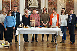 Das Foto zeigt mehrere Menschen in einer Reihe hinter einem Tisch stehen. Im Hintergrund ist die Ornamentverzierte Wand im Schloss Mannheim.