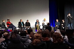 Das Foto zeigt mehrere Personen auf einer Bühne sitzen. Im Vordergrund sind viele Köpfe vom Publikum erkennbar.
