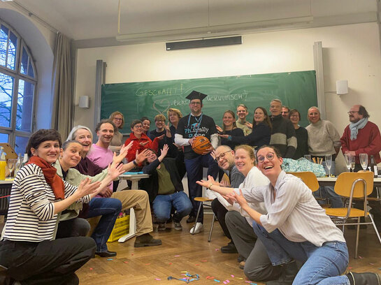 Das Bild zeigt David Dörrer, nach seiner erfolgreichen Disputation. Er steht in der Mitte eine Seminarraums, um ihn herum knien und stehen Freunde, Familienmitglieder und ein Teil des AW-ZIB-Teams. David Dörrer hält einen Basketball in der Hand und trägt auf dem Kopf einen Doktorhut.