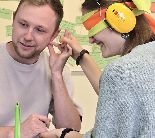 Eine Studentin hat die Augen verbunden und trägt auf den Ohren einen Gehörschutz. Sie erfährt, wie sich ein taubblinder Mensch fühlt. Sie wird dabei von einem Studenten begleitet.