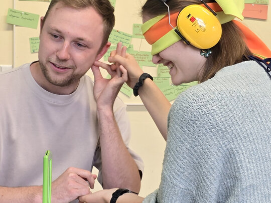 Eine Studentin hat die Augen verbunden und trägt auf den Ohren einen Gehörschutz. Sie erfährt, wie sich ein taubblinder Mensch fühlt. Sie wird dabei von einem Studenten begleitet.