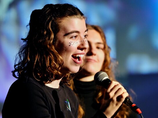 Das Foto zeigt zwei Personen auf der Bühne mit einem Mikrofon.