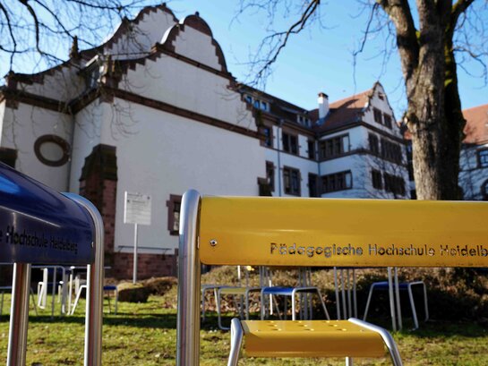 Die Außenaufnahme zeigt im Vordergrund zwei metallische Außenarbeotsplätze in gelb und blau mit der Inschrift "Pädagogische Hochschule Heidelberg". Im Hintergund ist ein Teil des Altbaus.