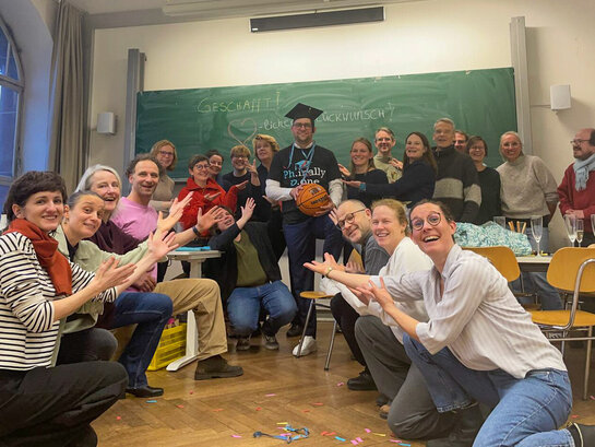 David Dörrer nach seiner erfolgreichen Disputation. Um ihn herum stehen und knien Freunde, Familienmitglieder und Mitglieder des AW-ZIB David Dörrer trägt einen Doktorhut und hält einen Basketball in der Hand.