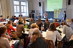 Das Bild zeigt einen Seminarraum, in dem eine Vielzahl von Studierenden sitzen. Ihr Blick ist nach vorne gerichtet, wo zwei Frauen eine Präsentation halten.