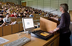 Auf dem Foto spricht eine Lehrende Person in einem Hörsaal zu den Studierenden.