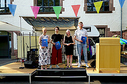 Auf dem Bild sind drei der vier Preisträger:innen zu sehen. Sie stehen gemeinsam mit Vera Heyl auf einer Bühne im Innenhof der Hochschule. Sie halten die Auszeichnungen in der Hand und schauen freundlich in die Kamera.