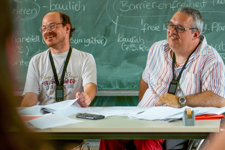 Das Bild zeigt die Bildungsfachkräfte Thilo Krahnke und Helmuth Pflantzer, wie sie ein Bildungsangebot ausbringen. Beide sitzen vor einer Tafel, auf der unter anderem die Worte "Barrierefreihet" zu lesen sind.