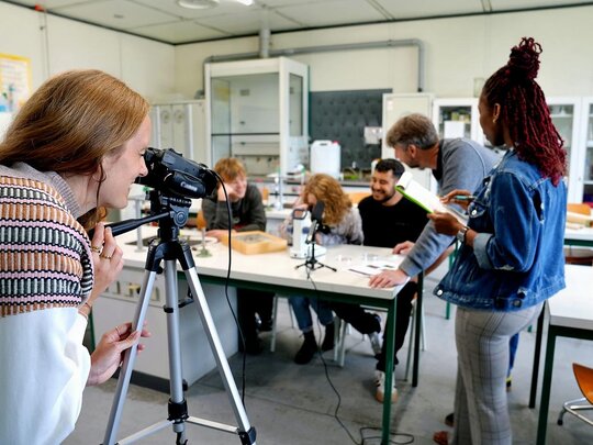 Der Fokus liegt auf einer Forscherin. Sie betrachtet eine Gruppe von Studierenden und einen Professor durch die Kamera. Das Bild wurde in einem Labor der Pädagogischen Hochschule Heidelberg aufgenommen. 