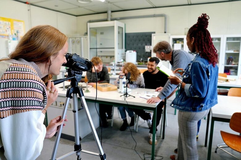 Der Fokus liegt auf einer Forscherin. Sie betrachtet eine Gruppe von Studierenden und einen Professor durch die Kamera. Das Bild wurde in einem Labor der Pädagogischen Hochschule Heidelberg aufgenommen. 