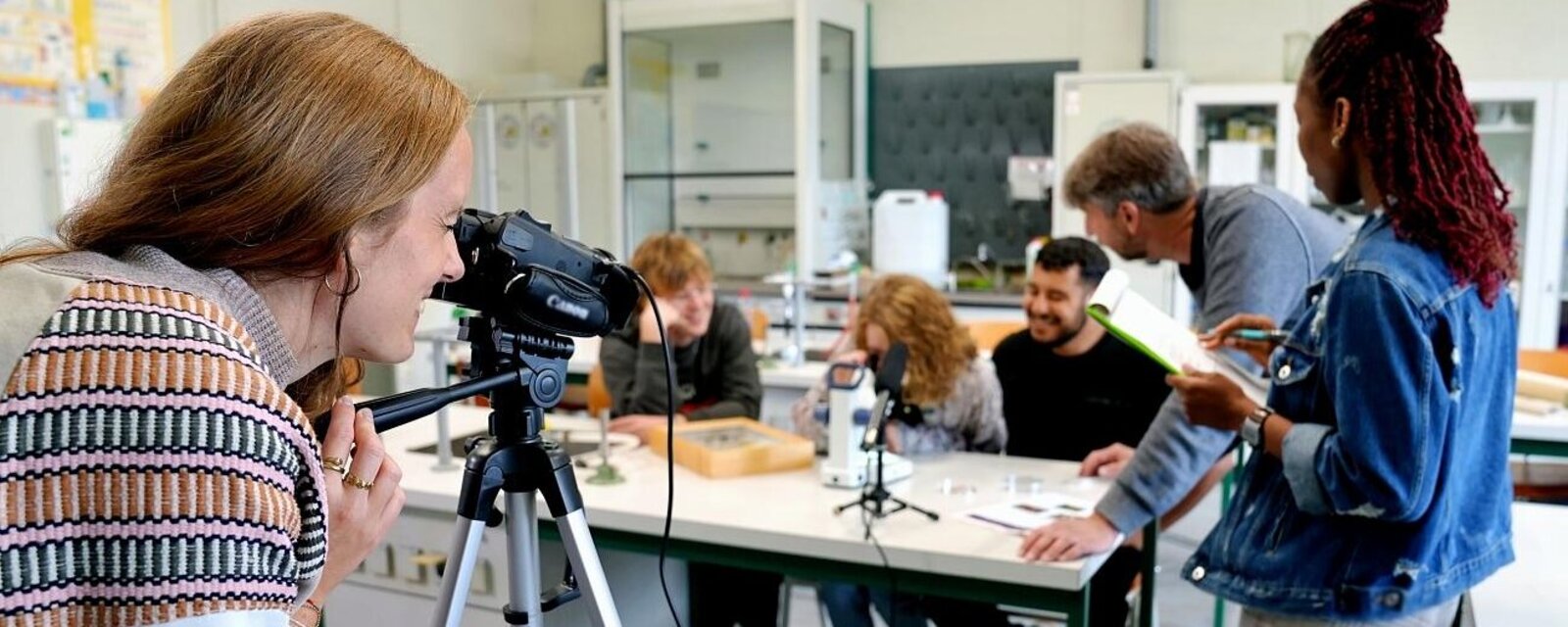Der Fokus liegt auf einer Forscherin. Sie betrachtet eine Gruppe von Studierenden und einen Professor durch die Kamera. Das Bild wurde in einem Labor der Pädagogischen Hochschule Heidelberg aufgenommen. 