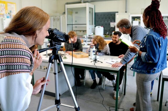 Der Fokus liegt auf einer Forscherin. Sie betrachtet eine Gruppe von Studierenden und einen Professor durch die Kamera. Das Bild wurde in einem Labor der Pädagogischen Hochschule Heidelberg aufgenommen. 