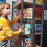 Auf dem Symbolbild sieht man ein junges Mädchen. Sie steht vor einem Bücherregal und greift hinein. Dabei schaut sie direkt in die Kamera. Sie trägt Corona-bedingt eine Mund-Nase-Schutz.