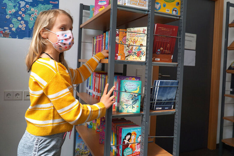 Auf dem Symbolbild sieht man ein junges Mädchen. Sie steht vor einem Bücherregal und greift hinein. Dabei schaut sie direkt in die Kamera. Sie trägt Corona-bedingt eine Mund-Nase-Schutz.