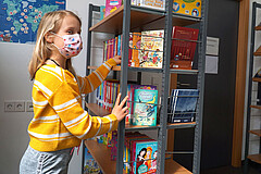 Auf dem Symbolbild sieht man ein junges Mädchen. Sie steht vor einem Bücherregal und greift hinein. Dabei schaut sie direkt in die Kamera. Sie trägt Corona-bedingt eine Mund-Nase-Schutz.