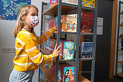 Auf dem Symbolbild sieht man ein junges Mädchen. Sie steht vor einem Bücherregal und greift hinein. Dabei schaut sie direkt in die Kamera. Sie trägt Corona-bedingt eine Mund-Nase-Schutz.