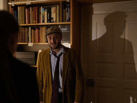 Das Foto zeigt eine Person vor einem Bücherregal in einem abgedunkelten Raum.