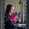 Das Symbolbild zeigt eine junge Frau im Tonstudio. Sie spricht lächelnd in ein Mikrofone.